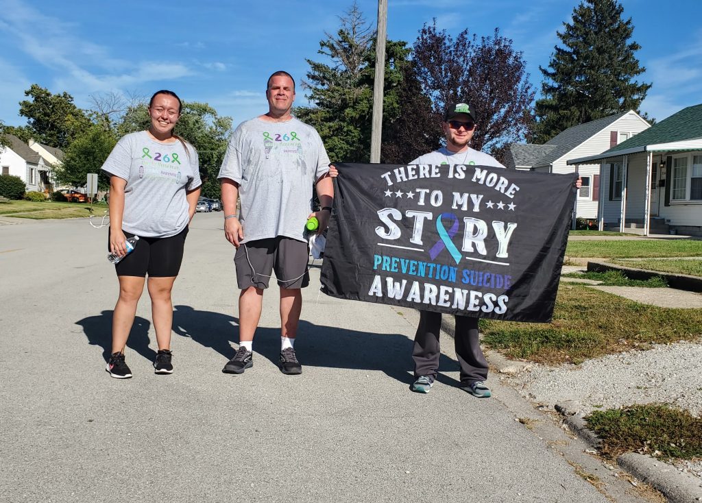 Amid high temperatures on Saturday, Jake Smith (holding banner) is spotted on Park Street in Manteno with teammates (lto r) Angela Urban - Manteno High, and Shaun Brav walking 26 miles to bring awareness and fundraising for mental health, and suicide. Amid honking car horns, supporters periodically joined them for a few miles and offered refreshments. Here they are seen at the 20 mile mark! Photo by Dan Gerber.