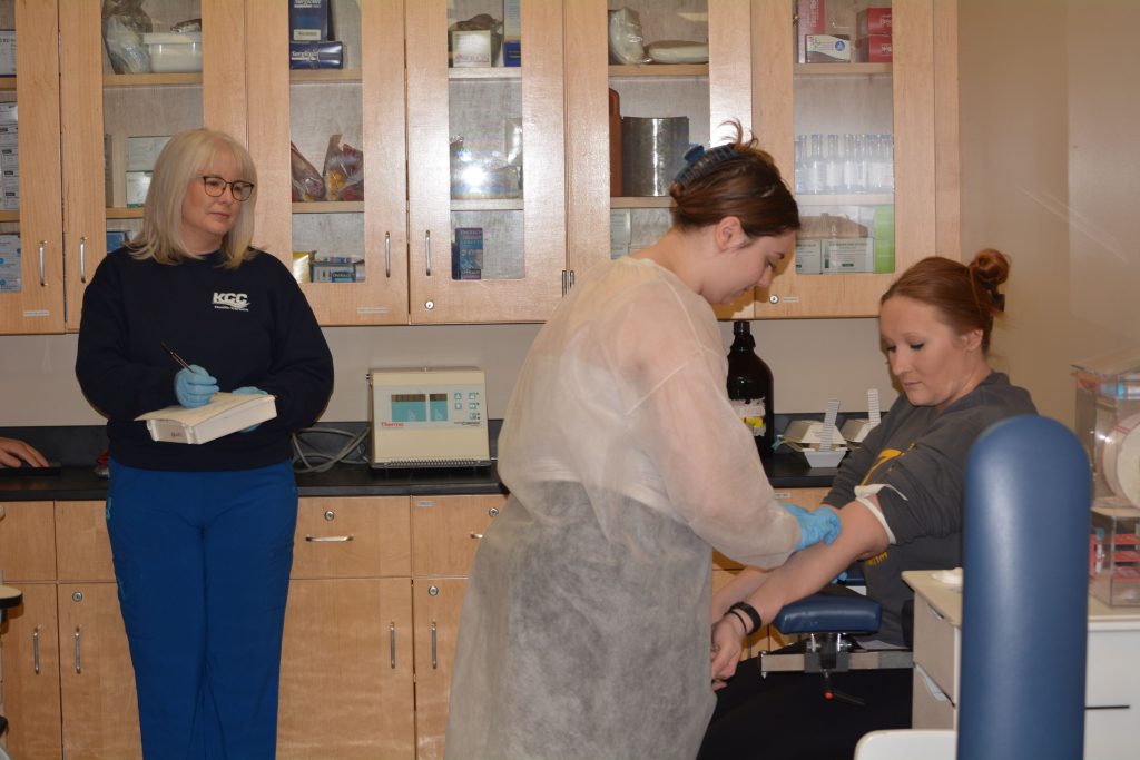 Roirie Bartucci draws blood from Dominique Schultz during a medical laboratory class at KCC, while, on left, is Lamanda Baade, professor and director of the Medical Laboratory Technology program. –Photo submitted.